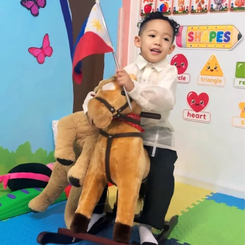 Happy preschooler in a barong riding a rocking horse and waving a Philippine flag beside the classroom shapes chart