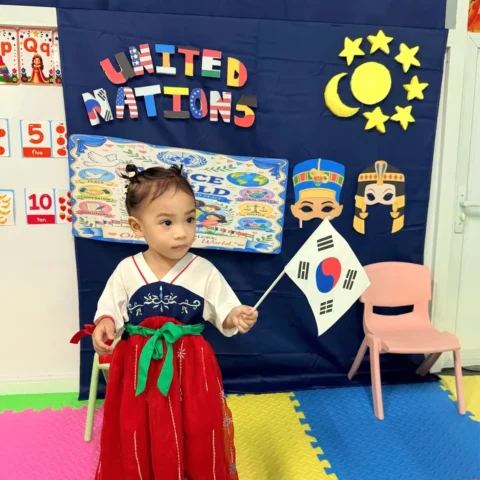 Preschooler in a traditional-style red and navy dress holding a South Korean flag in front of the United Nations Day display