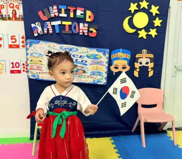 Preschooler in a traditional-style red and navy dress holding a South Korean flag in front of the United Nations Day display