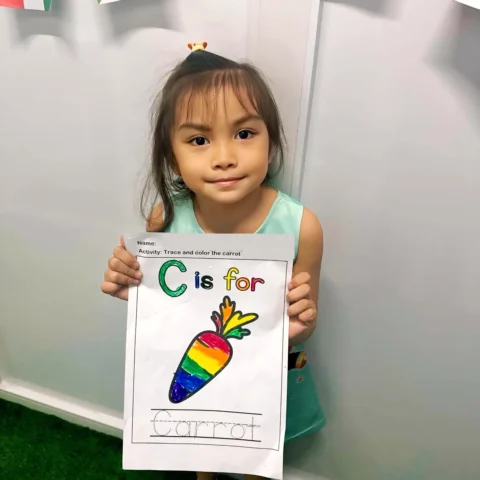 Smiling preschooler proudly holding up her rainbow-coloured 'C is for carrot' worksheet