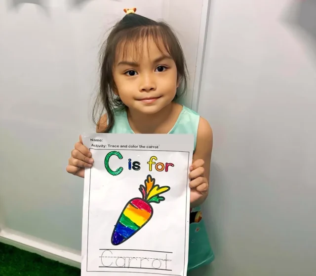 Smiling preschooler proudly holding up her rainbow-coloured 'C is for carrot' worksheet