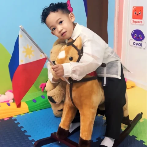 Toddler in a barong Tagalog waving a Philippine flag on a rocking horse in front of the classroom's painted rainbow mural