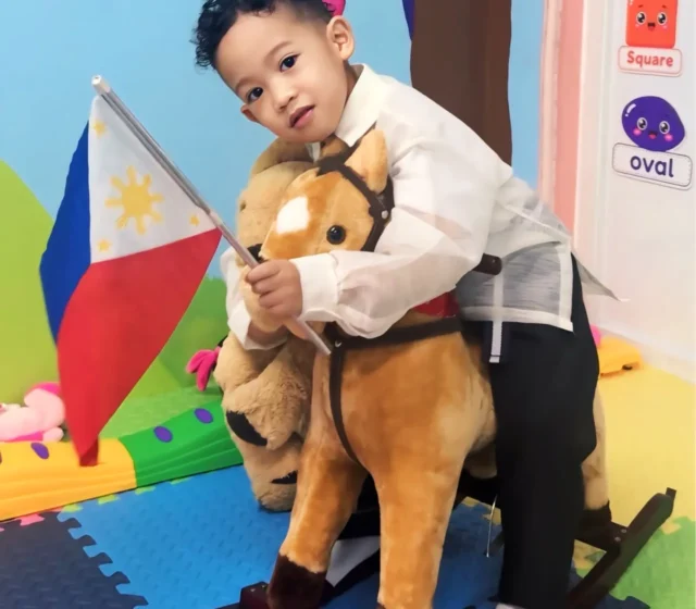 Toddler in a barong Tagalog waving a Philippine flag on a rocking horse in front of the classroom's painted rainbow mural