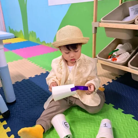 Toddler in a straw hat sitting on foam mats and exploring numbered paper cups during a counting activity