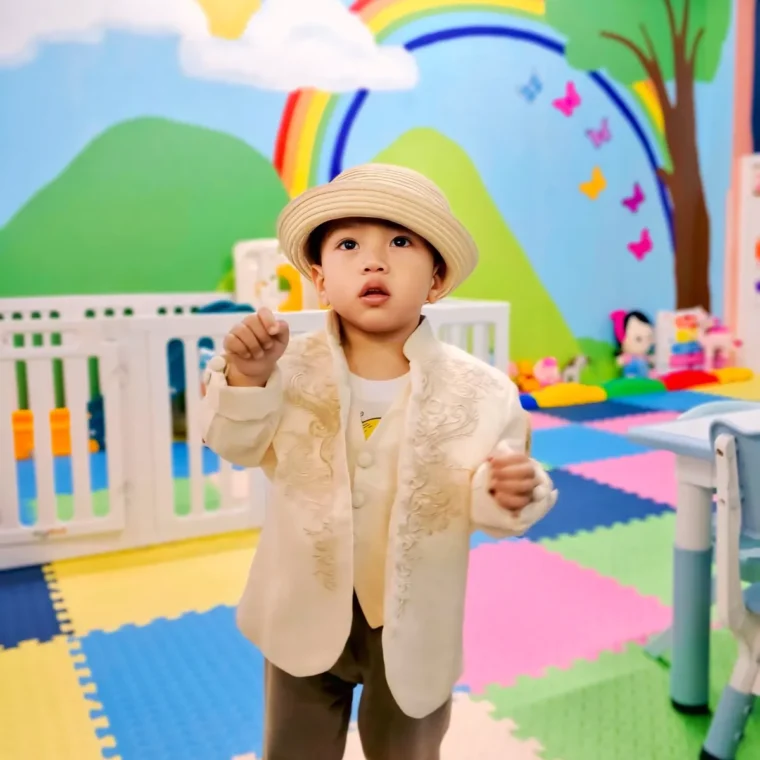 Toddler in an embroidered cream jacket and straw hat standing in the bright playroom with a painted rainbow and hills behind
