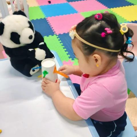 Little girl fitting a craft stick into a paper cup at a classroom table beside a panda plush, with colourful foam mats behind