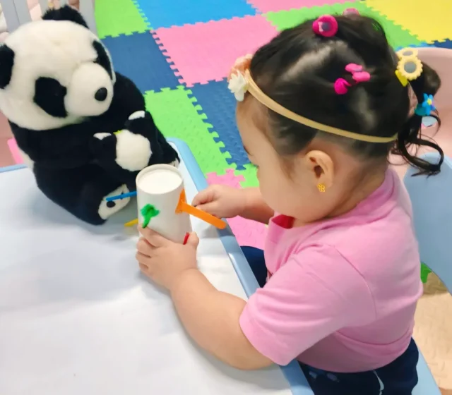 Little girl fitting a craft stick into a paper cup at a classroom table beside a panda plush, with colourful foam mats behind