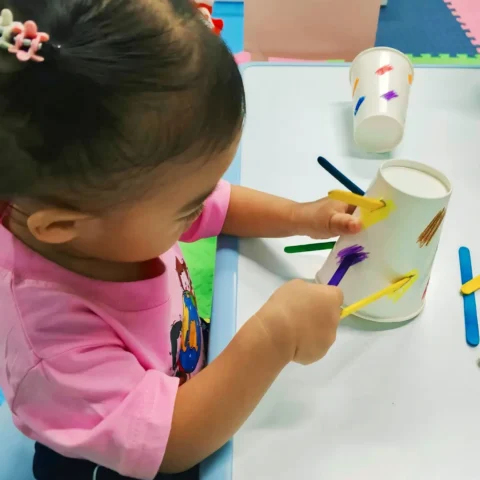 Toddler pushing coloured craft sticks into the slots of a paper cup during a fine-motor activity