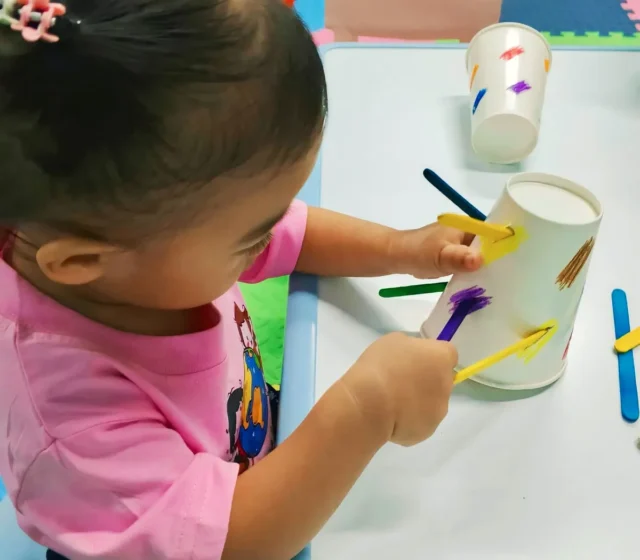 Toddler pushing coloured craft sticks into the slots of a paper cup during a fine-motor activity