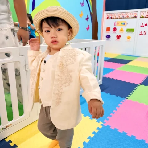 Toddler in a straw hat and embroidered jacket walking across the colourful foam-mat playroom, with alphabet and shapes charts behind