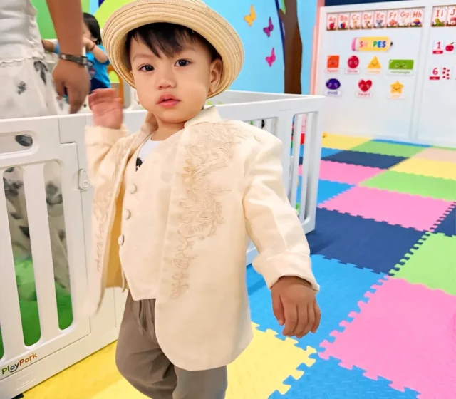 Toddler in a straw hat and embroidered jacket walking across the colourful foam-mat playroom, with alphabet and shapes charts behind
