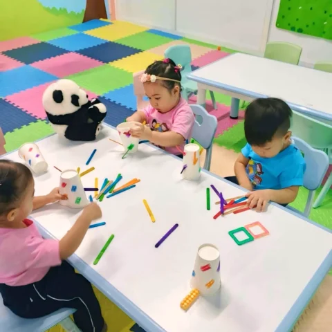 Three toddlers at a low classroom table sorting colourful craft sticks into paper cups, with shapes and numbers charts on the wall