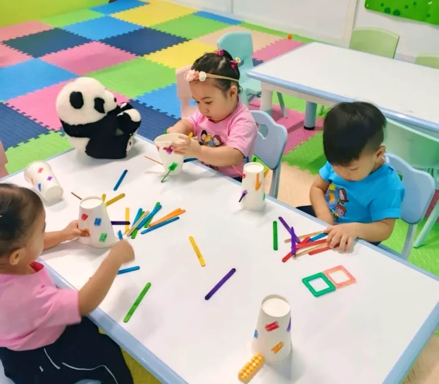Three toddlers at a low classroom table sorting colourful craft sticks into paper cups, with shapes and numbers charts on the wall