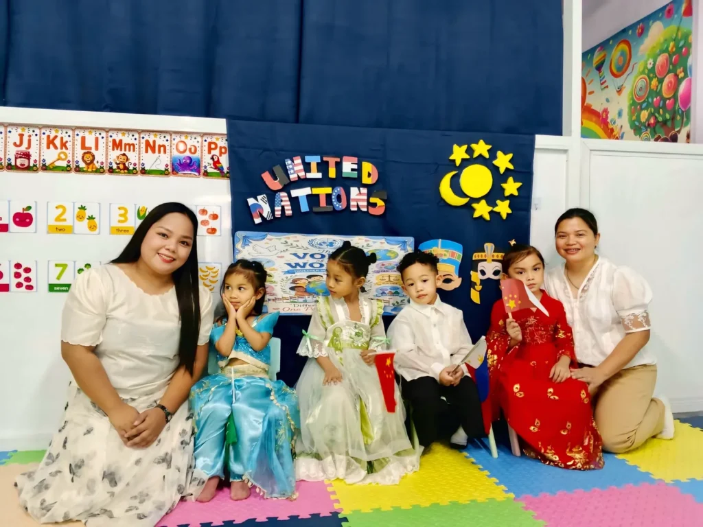 Two smiling teachers kneeling with preschoolers dressed in costumes from different countries at the United Nations Day celebration