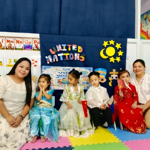 Two smiling teachers kneeling with preschoolers dressed in costumes from different countries at the United Nations Day celebration
