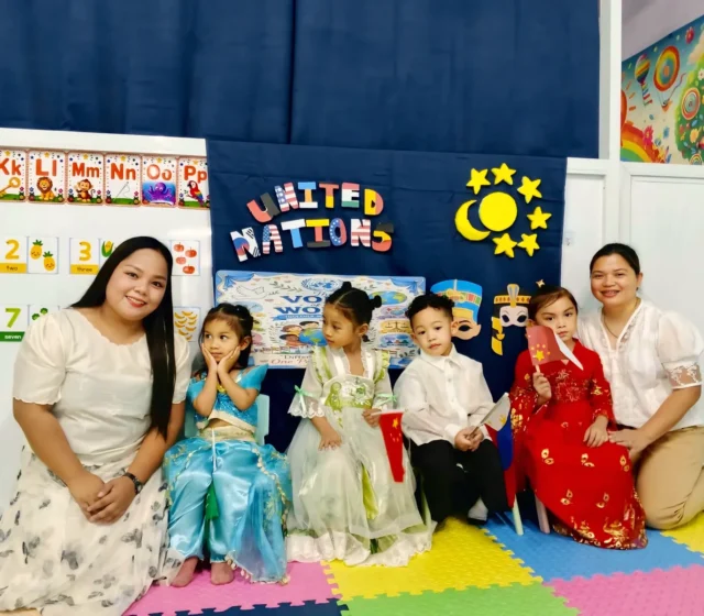 Two smiling teachers kneeling with preschoolers dressed in costumes from different countries at the United Nations Day celebration